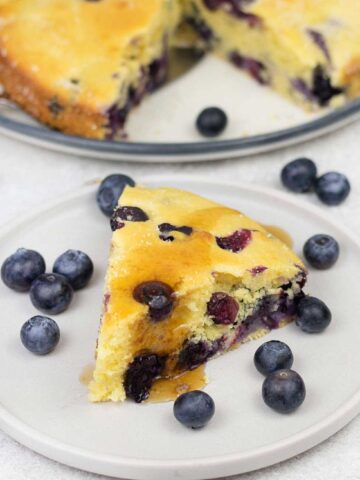 A slice of the blueberry cornbread topped with maple syrup and some blueberries are in the plate.