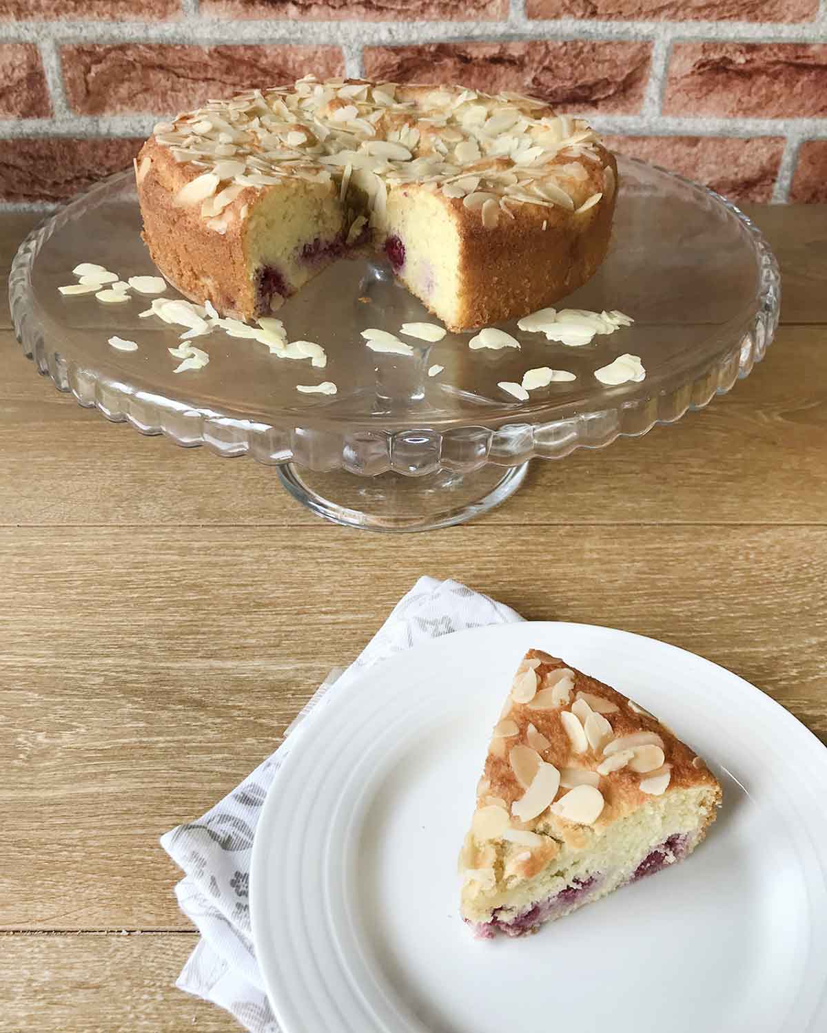 Almond raspberry cake on a cake stand and a slice is on a plate.