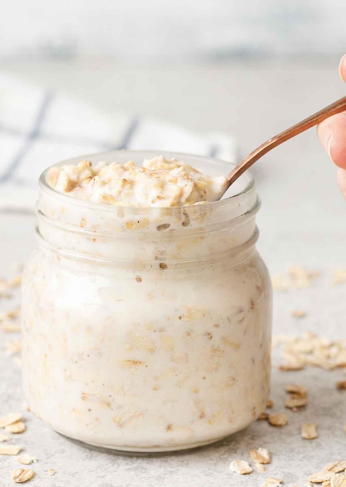 Taking a spoonful of basic overnight oats from a glass jar and some rolled oats in the table.