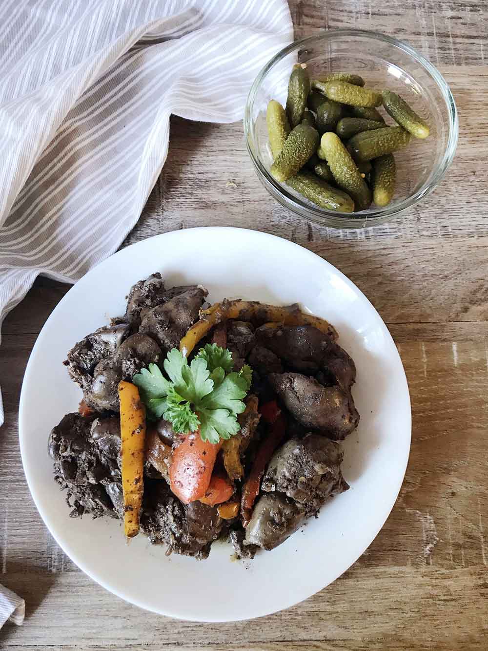 A plate full of pan-fried chicken livers with onion and pepper and a bowl of gherkins.