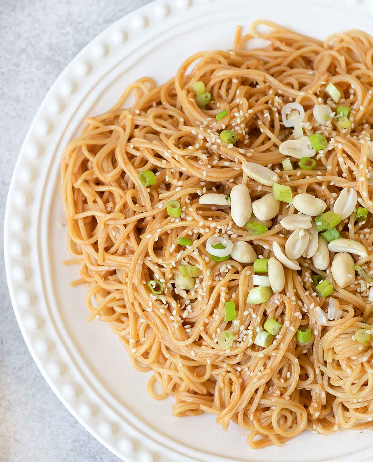 Peanut sesame noodles in a white plate topped with sesame seeds and some peanuts.