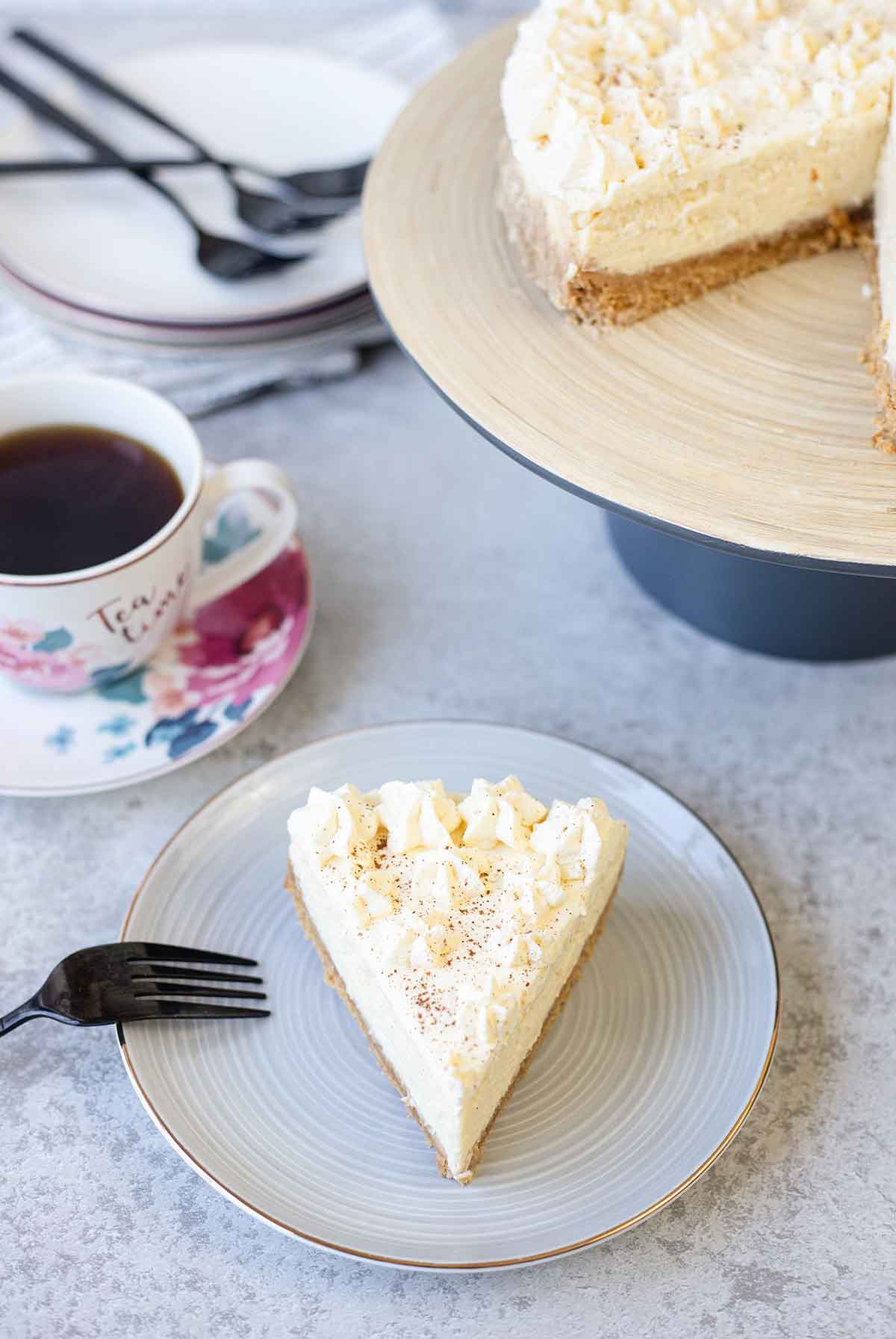A slice of the eggnog cheesecake is in a plate and a cup of tea next to it.